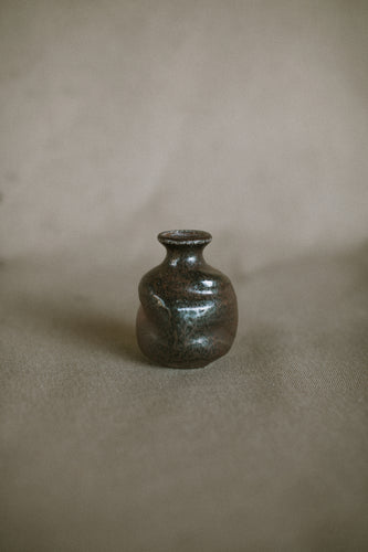 A small brown and black bud vase on a beige fabric background. The piece is made from Iron rich Australian stoneware clay wood ash covering one side.