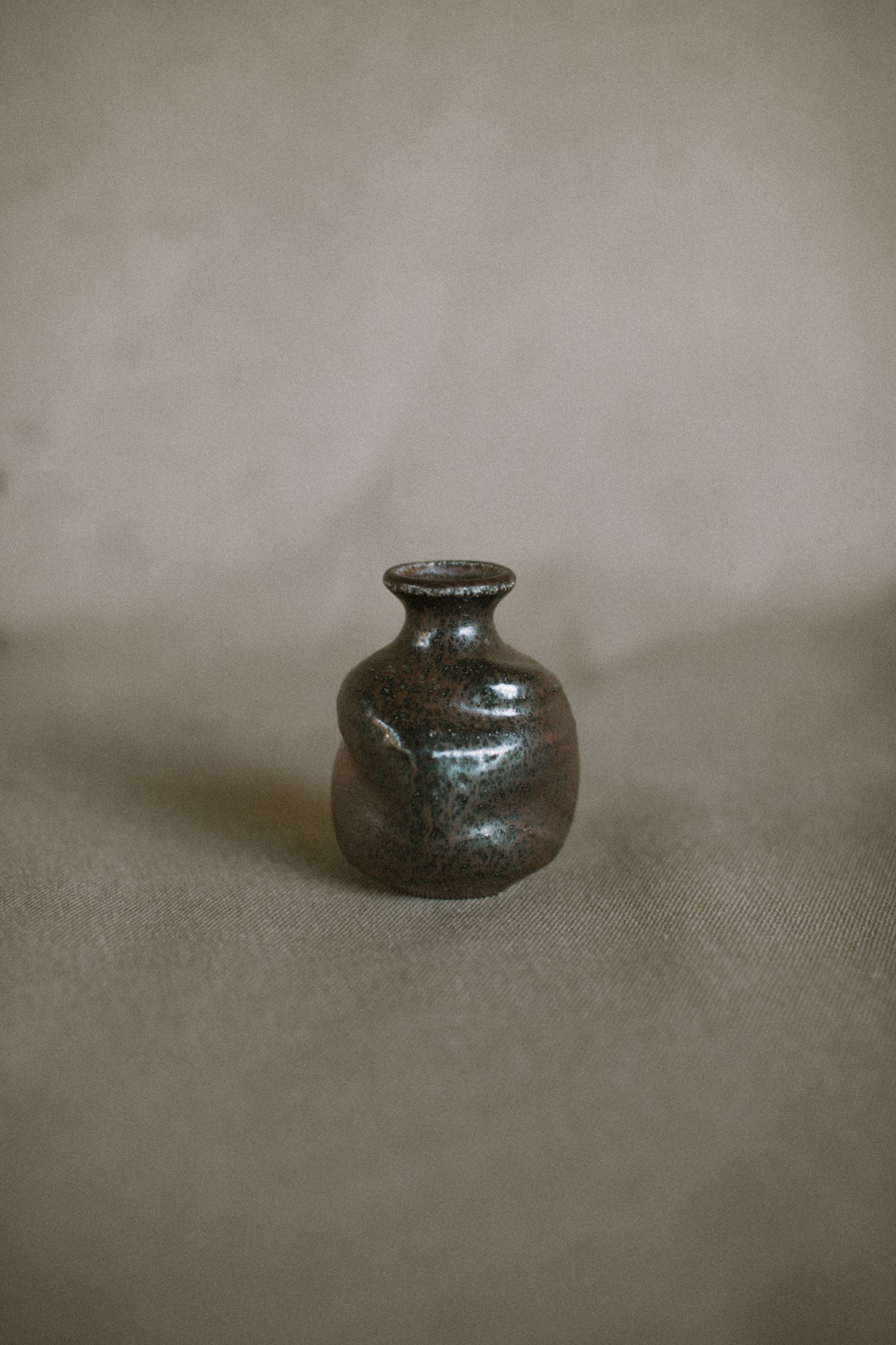 A small brown and black bud vase on a beige fabric background. The piece is made from Iron rich Australian stoneware clay wood ash covering one side.