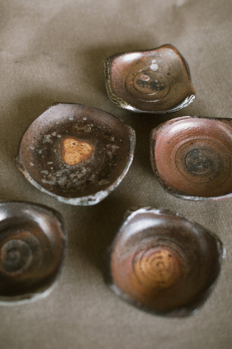 A collection of wood fired trinket dishes on a beige fabric background. These wheel thrown small plates are made from Iron rich Australian stoneware clay with a variety of brown, orange and black markings.