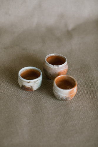 A collection of small wood fired ceramic shot glasses on a beige fabric background. These wheel thrown pieces are made from white Australian stoneware clay that has an orange Shino glaze and dark brown Iron brush work.