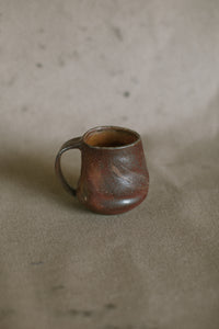 A dark brown mug on a beige fabric background. This piece is made from iron rich Australian stoneware clay that has turned an almost metallic brown in the wood kiln. Inside is a light orange Shino glaze