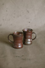 Load image into Gallery viewer, Two dark brown wood fired ceramic mugs on a beige fabric background. These wheel thrown pieces are made from iron rich Australian stoneware clay that has turned an almost metallic brown in the wood kiln. Inside is a reddy brown Shino glaze.