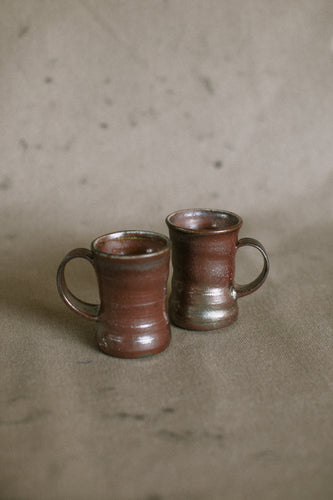 Two dark brown wood fired ceramic mugs on a beige fabric background. These wheel thrown pieces are made from iron rich Australian stoneware clay that has turned an almost metallic brown in the wood kiln. Inside is a reddy brown Shino glaze.