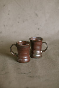 Two dark brown wood fired ceramic mugs on a beige fabric background. These wheel thrown pieces are made from iron rich Australian stoneware clay that has turned an almost metallic brown in the wood kiln. Inside is a reddy brown Shino glaze.