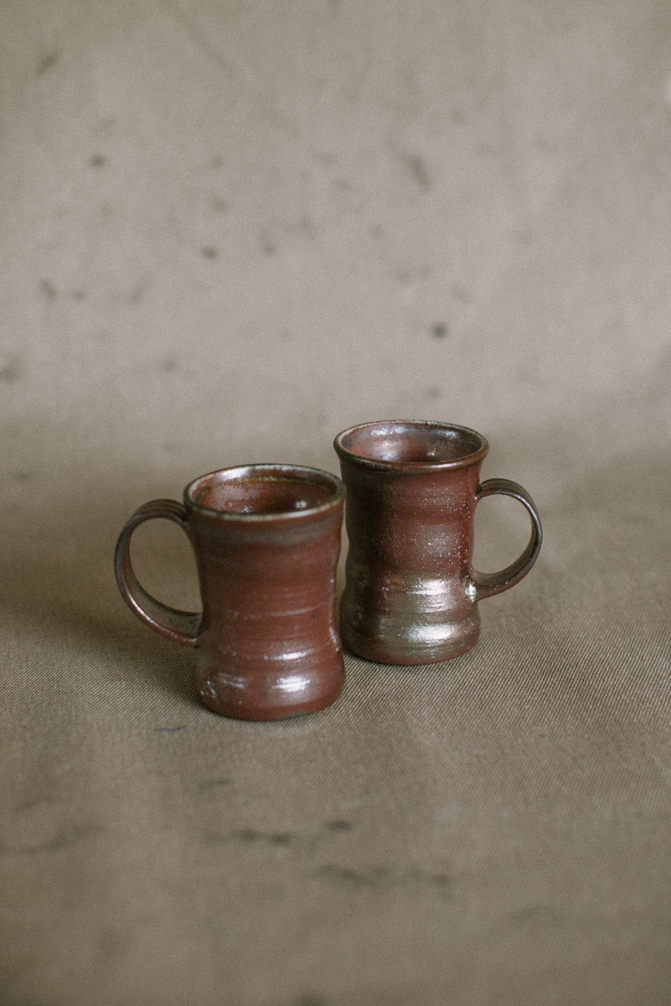Two dark brown wood fired ceramic mugs on a beige fabric background. These wheel thrown pieces are made from iron rich Australian stoneware clay that has turned an almost metallic brown in the wood kiln. Inside is a reddy brown Shino glaze.