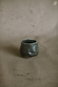 A small dark black blue wood fired cup on a beige fabric background. This wheel thrown and altered Japanese inspired yunomi is made from dark speckled Australian stoneware clay that has a Cobalt blue glaze that has been reduction fired in a wood kiln