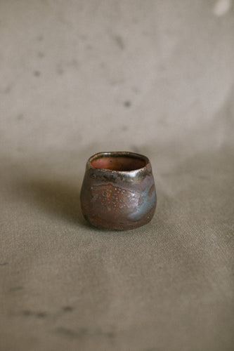 A small dark brown wood fired cup on a beige fabric background. This wheel thrown and altered Japanese inspired yunomi is made from dark Iron rich Australian stoneware clay that has a Shino glaze inside.
