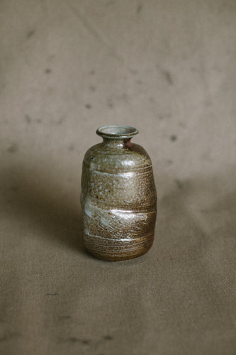 A dark brown wood fired vase covered in green ash on a beige fabric background. This wheel thrown and altered Japanese inspired flower vase is made from dark Iron rich Australian stoneware clay with a white slip and Sally wattle ash.