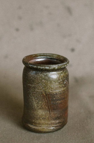 A dark brown wood fired vase on a beige fabric background. This wheel thrown and altered Japanese inspired flower vase is made from dark Iron rich Australian stoneware clay with Sally wattle ash on one side.