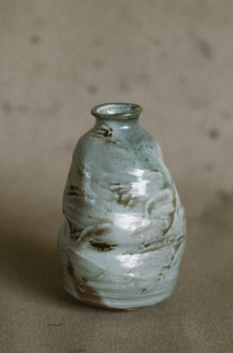 A grey and brown wood fired vase on a beige fabric background. This wheel thrown and altered flower vase is made from white Australian stoneware clay with Iron brush work and light wood ash covering the piece.