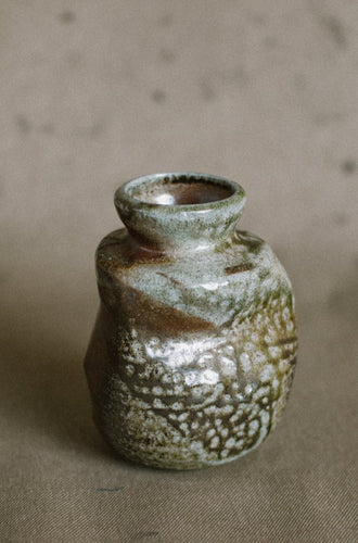 A green and brown wood fired vase on a beige fabric background. This wheel thrown and altered Japanese inspired vase is made from Iron rich Australian stoneware clay with dark green Sally wattle ash on one side.