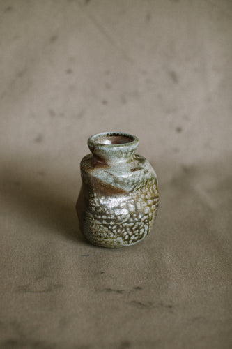 A green and brown wood fired vase on a beige fabric background. This wheel thrown and altered Japanese inspired vase is made from Iron rich Australian stoneware clay with dark green Sally wattle ash on one side.