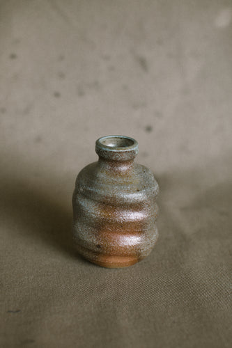 A green and brown wood fired vase on a beige fabric background. This wheel thrown and altered flower vase is made from sandy orange Australian stoneware clay with Iight green wood ash.