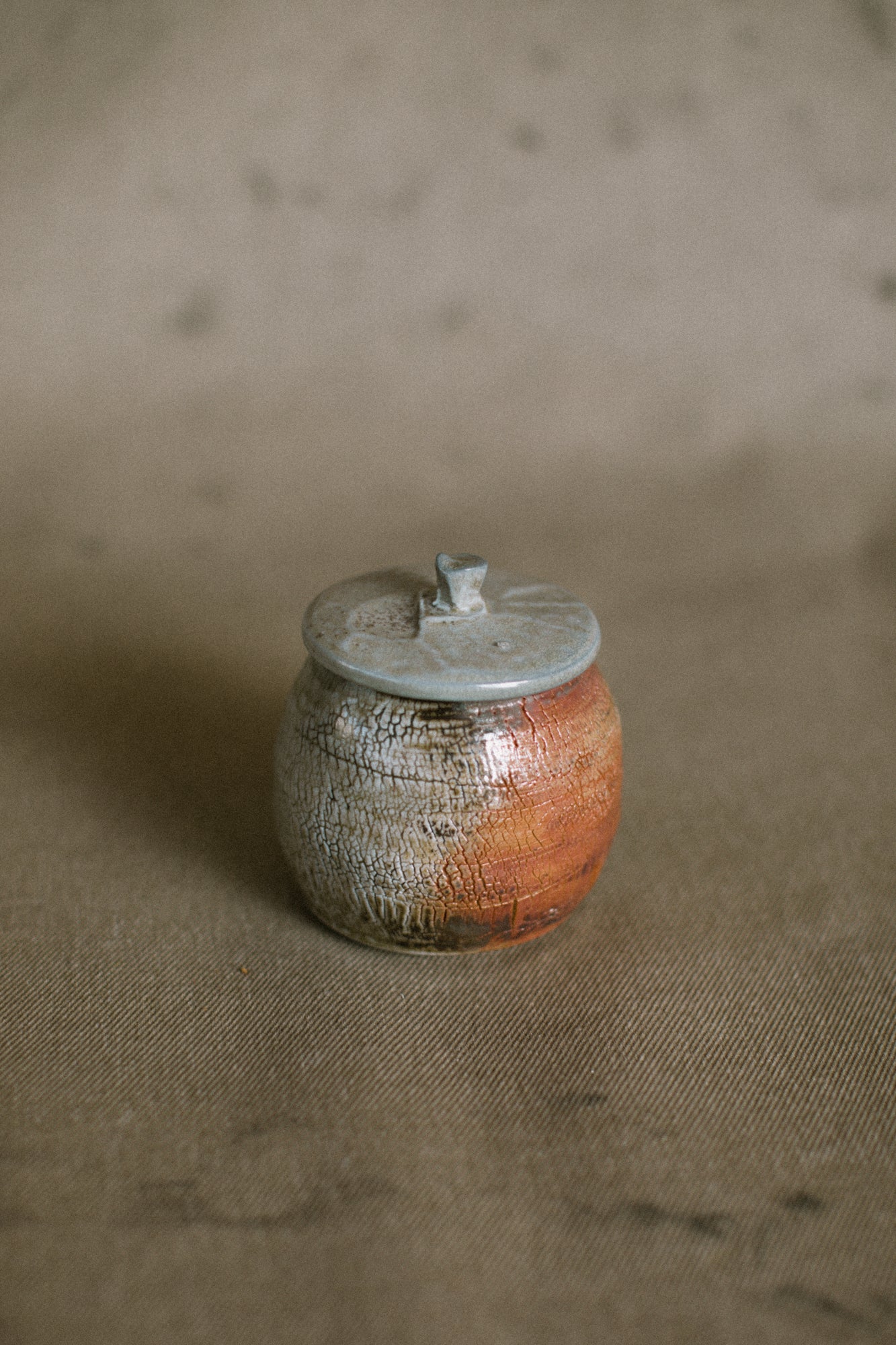 A grey and orange wood fired jar on a beige fabric background. This wheel thrown jar is made from white Australian stoneware clay with an orange wild clay slip covered in light green wood ash.