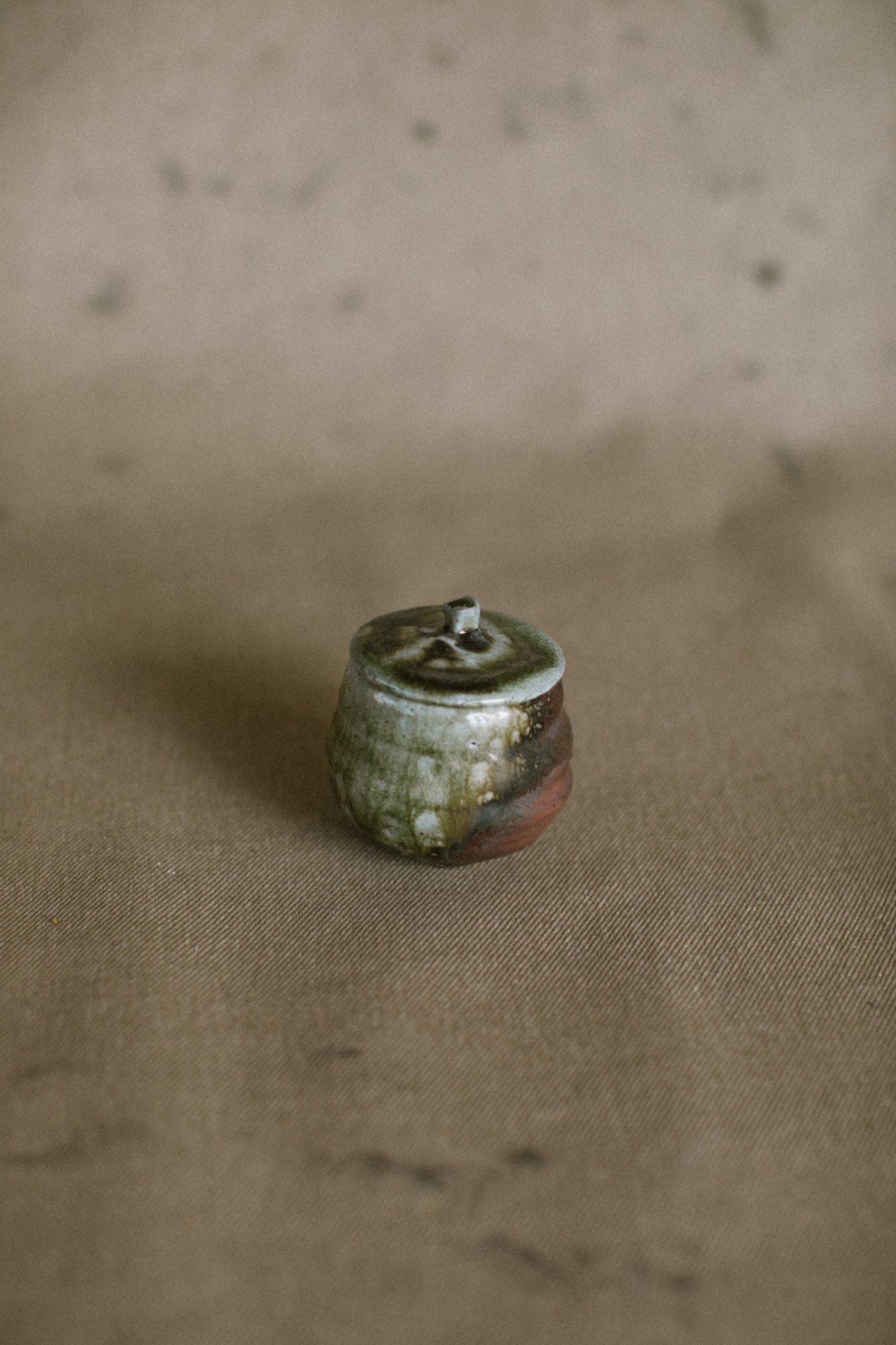 A brown and green wood fired jar on a beige fabric background. This wheel thrown jar is made from an Iron rich Australian stoneware clay with dark green wood ash covering one side.