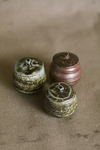 Three brown and green small wood fired jars on a beige fabric background. These wheel thrown jars are made from iron rich Australian stoneware clay with dark green heavy wood ash covering two
