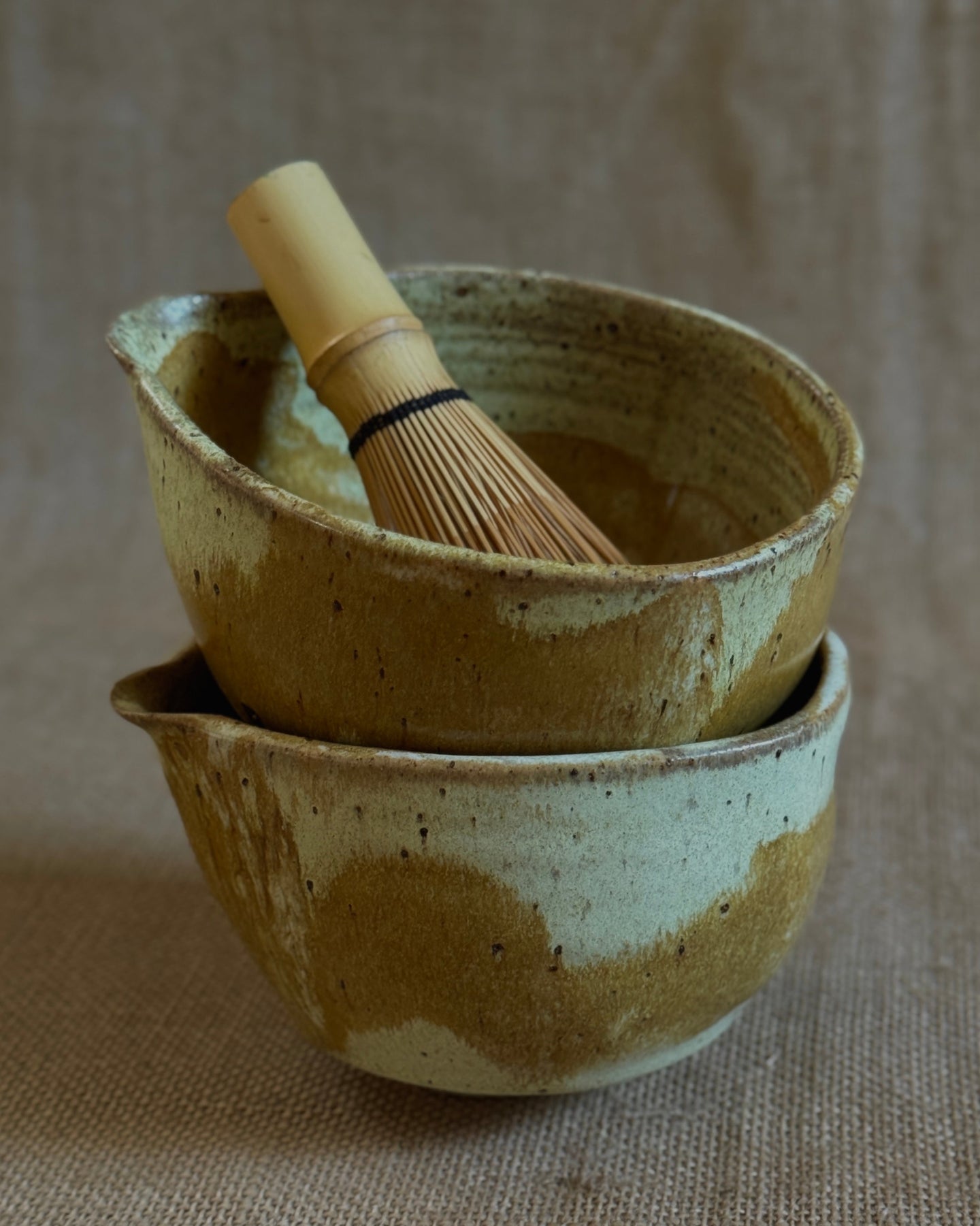 Two ceramic matcha bowls with a chasen (bamboo whisk) on a textured beige background