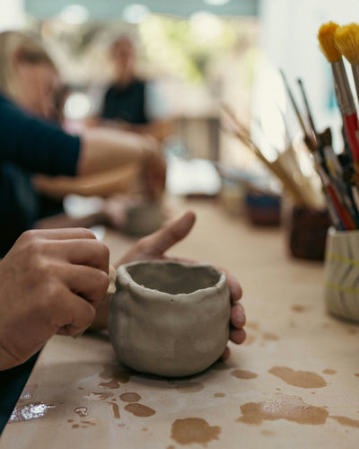 A person holding a clay pinch pot and stamping detail into it. The ceramic cup is sitting on a table in a pottery studio with other people working on clay projects in the background